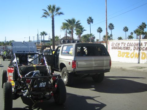 1990 Ford Bronco - tan lifted pickup with a bed shell tows a tube-frame off-road buggy numbered 1613 on a palm-lined street.