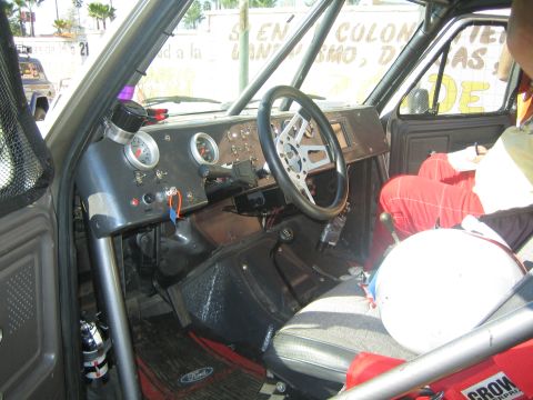 1990 Ford Bronco - stripped interior of an off-road race vehicle shows a roll cage, round gauges, a worn steering wheel and a red seat.