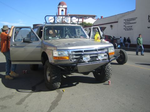 1990 Ford Bronco - silver lifted pickup with a roof light bar, brush guard and oversized tires is parked on a street by a domed building.