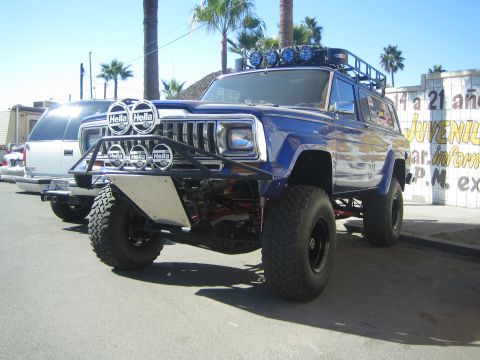 1990 Ford Bronco - blue lifted full-size SUV with stacked Hella lights, a tube brush guard and big mud tires parked on a street.