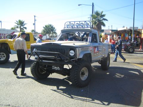 1990 Ford Bronco - white lifted off-road pickup with a roof light bar and brush guard sits on a busy street as people walk nearby.