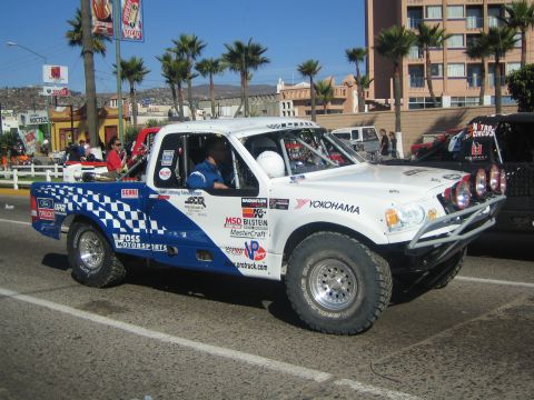 1990 Ford Bronco - white off-road race truck covered in Yokohama, MSD and other sponsor decals drives a street lined with palms and hotels.