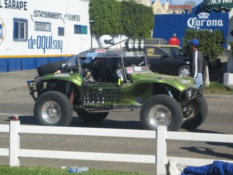1990 Ford Bronco - green tube-frame off-road buggy with a helmeted driver waits on a street in front of a blue wall and Corona sign.