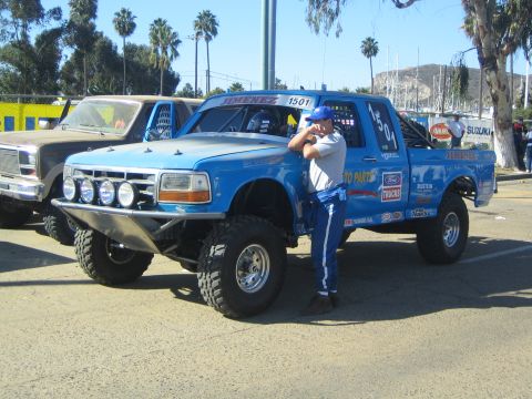 1990 Ford Bronco - man in a blue racing suit leans against a lifted blue Ford pre-runner race truck with a roof-mounted light bar at an off-road event.