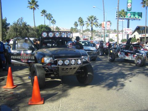 1990 Ford Bronco - black off-road pre-runner race truck with a roof light bar and spare tire sits on a palm-lined street behind orange traffic cones.