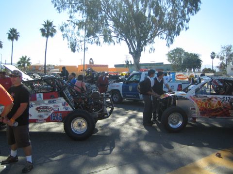 1990 Ford Bronco - lineup of off-road race buggies and trucks, including number 645, parked along a sunny palm-lined street with spectators and police nearby.