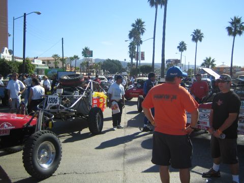 1990 Ford Bronco - Spectators and crew gather around off-road race buggies, including number 1206, parked on a palm-lined street under a clear blue sky.