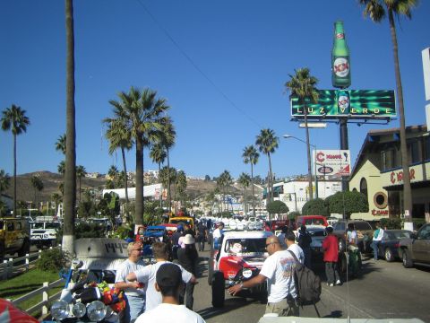 1990 Ford Bronco - busy street lined with palm trees and crowds, with off-road race vehicles and a large XX/Luz Verde beer billboard overhead.