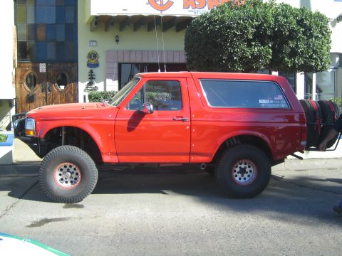 1990 Ford Bronco - Side profile of a red full-size Ford Bronco lifted on oversized tires with rear-mounted spare tires, parked in front of a restaurant.