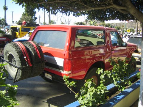 1990 Ford Bronco - Rear three-quarter view of a red full-size Ford Bronco with rear-mounted dual spare tires, parked behind a planter of green ivy.