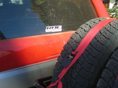 1990 Ford Bronco - Close-up of a red SUV's rear corner showing an 'RDC' window decal and a spare tire secured by a red ratchet strap.