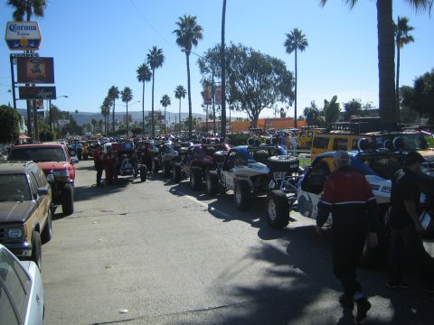 1990 Ford Bronco - long line of off-road race trucks and buggies staged along a sunny palm-lined street, with a Corona beer sign at left.