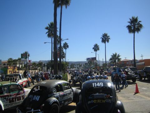 1990 Ford Bronco - Two off-road racing Volkswagen Beetles numbered 1148 and 1149 staged among other race cars on a sunny palm-lined street.
