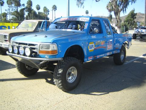 1990 Ford Bronco - blue Ford pre-runner race truck number 1501 with a roof light bar and NAPA, Ford and Auto Parts sponsor decals at a staging area.