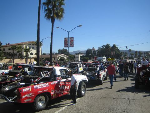 1990 Ford Bronco - Off-road race trucks, including a red number 233, staged along a street with spectators and a Spanish-style building in the background.