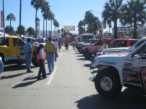 1990 Ford Bronco - Rows of off-road race trucks and buggies, including Yokohama-sponsored entries, staged with spectators on a palm-lined street.