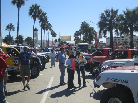 1990 Ford Bronco - Staged off-road race trucks and buggies line a palm-lined boulevard as officials in safety vests and spectators gather among them.