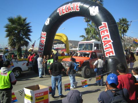 1990 Ford Bronco - orange off-road truck passes under a black inflatable Tecate start/finish arch as a crowd of spectators watches.