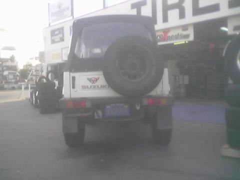 1990 Ford Bronco - Rear of a white Suzuki Samurai showing the tailgate-mounted spare and a license plate, parked at a shop.