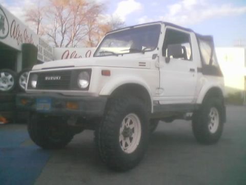 1990 Ford Bronco - white lifted Suzuki Samurai with chrome wheels and bare trees behind it parked near an Alfa Italia sign.
