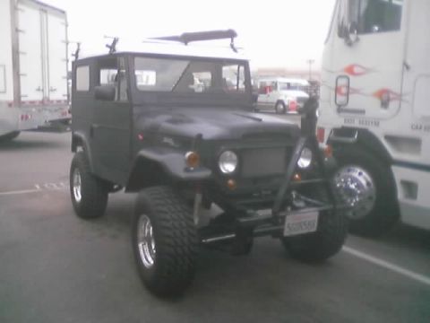 1990 Ford Bronco - gray lifted vintage hardtop 4x4 with round headlights and chrome wheels parked between semi trucks and trailers.