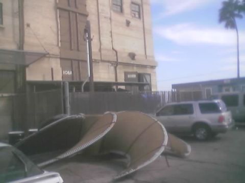 1990 Ford Bronco - silver SUV sits in a lot beside a tall aged industrial building with curled sheet-metal debris on the ground.