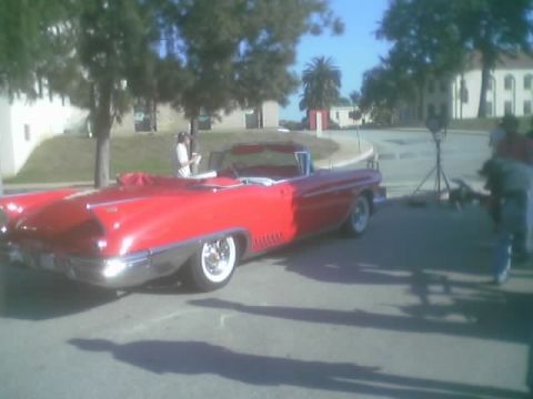 1990 Ford Bronco - Rear quarter of a red Cadillac convertible with tailfins parked on pavement near trees and buildings.