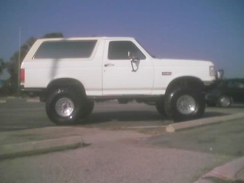 1990 Ford Bronco - white lifted full-size Ford Bronco SUV on oversized tires parked in an empty lot under blue sky.