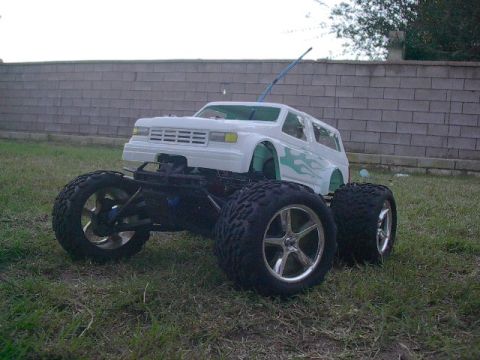 1993 Ford Bronco - A white remote-controlled monster truck with teal flame decals sits on a grassy lawn in front of a brick wall.