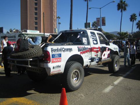 1990 Ford Bronco - white Ford off-road race truck numbered 462, decked with sponsor decals and a tailgate spare, parked on a sunny palm-lined street.