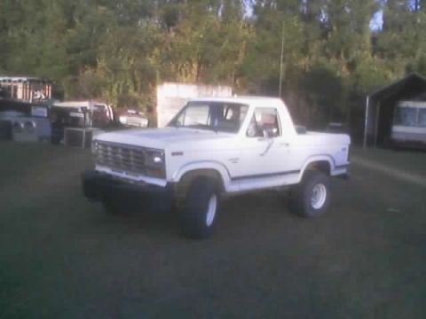 1986 Ford Bronco - This lifted 1986 Ford Bronco is shown in a three-quarter front view, parked on a grassy area with trees in the background.