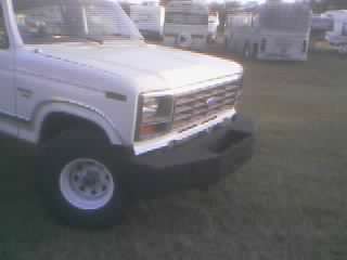 1986 Ford Bronco - Close-up of the front of a white SUV, showing its grille and bumper. Other vehicles are visible in the distance.