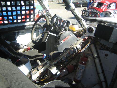 1990 Ford Bronco - gutted cockpit of an off-road race truck showing a steering wheel, gauge cluster, roll cage tubing and a red fire extinguisher.
