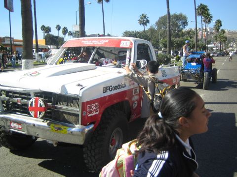 1990 Ford Bronco - white off-road race truck numbered 305 idles on a palm-lined street while a schoolgirl with a backpack walks by in the foreground.