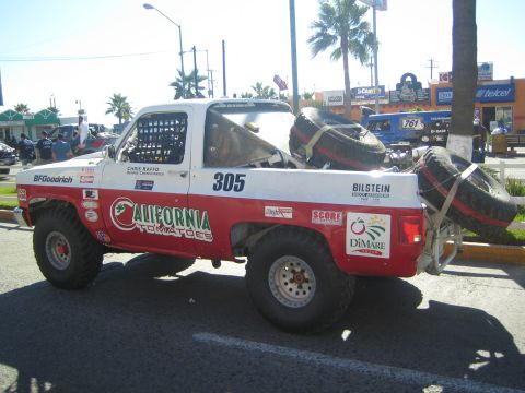 1990 Ford Bronco - red and white #305 off-road race truck lettered California Tomatoes parked along a busy commercial street with palm trees.