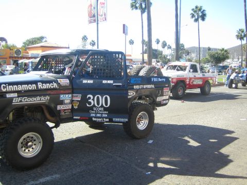 1990 Ford Bronco - black #300 race truck lettered Cinnamon Ranch and a white #305 truck staged on a palm-lined street near vendor stands.