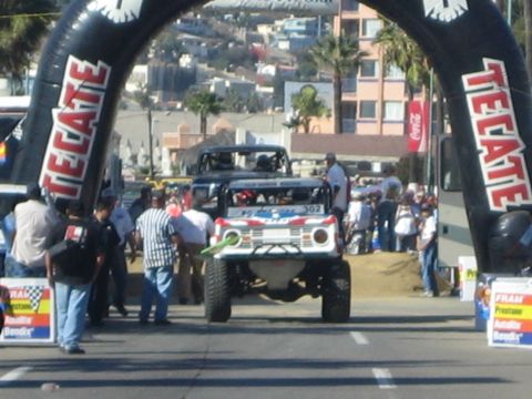 1990 Ford Bronco - off-road race truck rolls under an inflatable Tecate arch on a street lined with spectators, banners and a coastal hillside backdrop.