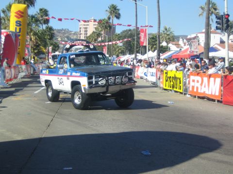 1990 Ford Bronco - blue and white #349 off-road race truck drives down a street flanked by spectators and FRAM and Suzuki banners under palm trees.