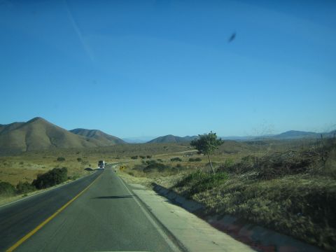1990 Ford Bronco - two-lane highway curves through arid brown hills and scrub under a deep blue sky, with a truck in the distance.