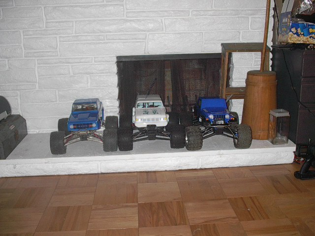 Three RC monster trucks lined up on a fireplace hearth. One is blue, one is white, and one is blue