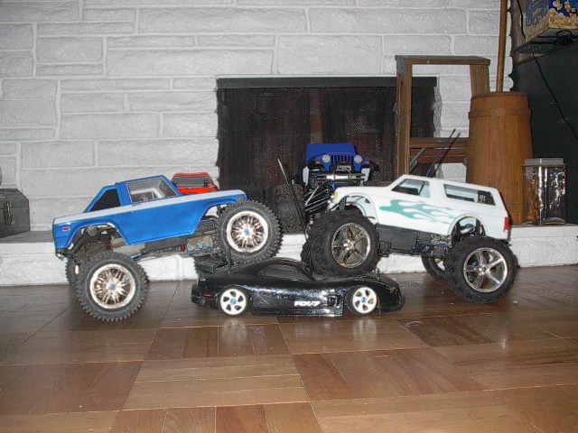 RC monster trucks, one blue and white, one white with flames, stacked on a black RC sports car. A blue RC Jeep is behind them