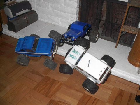 1993 Ford Bronco - Three remote control monster trucks are positioned on a wooden floor near a fireplace.
