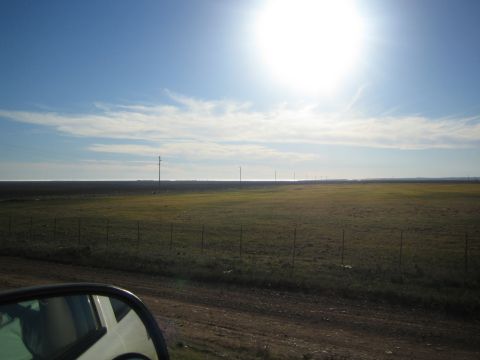 1990 Ford Bronco - Flat grassland with a wire fence and power poles seen past a car side mirror, the sun glaring high in a pale blue sky.