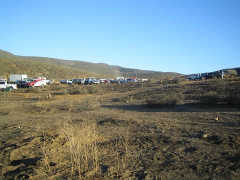 1990 Ford Bronco - row of parked trucks, RVs and trailers gathered in a barren brown desert valley ringed by low hills under a clear blue sky.