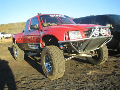 1990 Ford Bronco - red Ford Ranger extended-cab prerunner truck with a tubular front bumper, auxiliary lights, and a long-travel suspension on dirt.