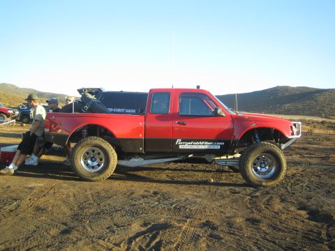 1990 Ford Bronco - Side profile of a two-tone red and black Ford Ranger prerunner with fender flares parked on a dirt lot as people stand nearby.