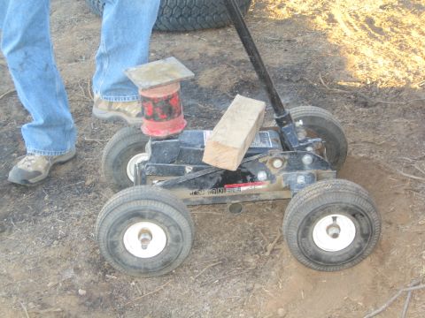 1990 Ford Bronco - black hydraulic floor jack topped with a wood block and red object, positioned beside a person's legs on dusty ground.
