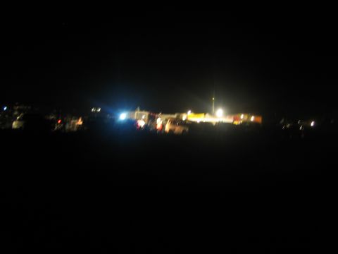 1990 Ford Bronco - blurry nighttime scene showing distant lights, tents, and vehicles glowing across a dark desert landscape.