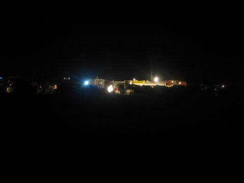 1990 Ford Bronco - Floodlit tents and a yellow banner glow in the distance across a dark desert at night.