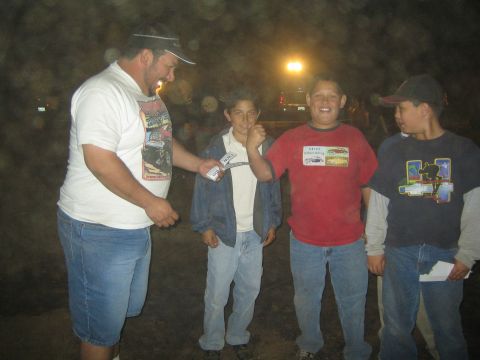 1990 Ford Bronco - heavyset man hands out cards to three smiling boys standing on dirt under a streetlight at night.